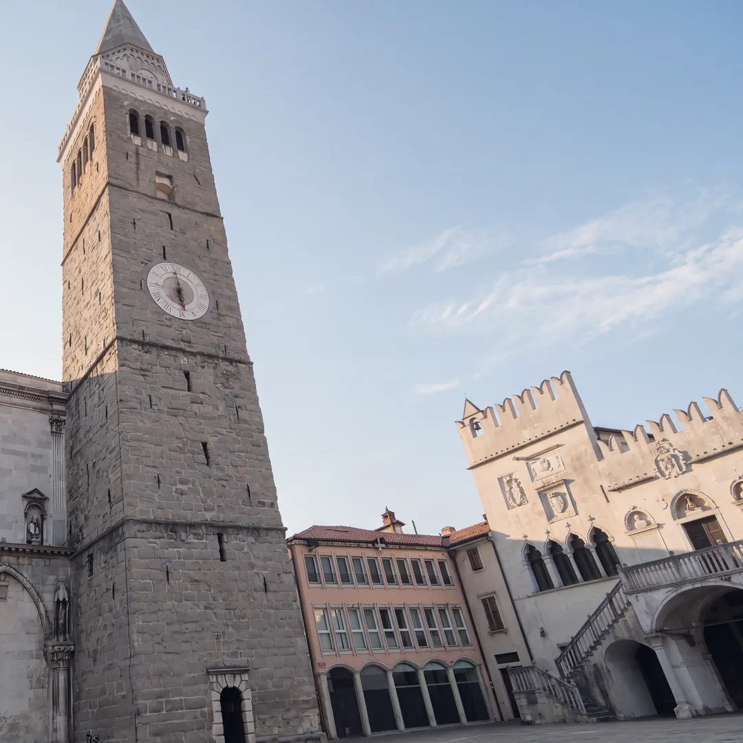 Tito Square in Koper at first light, empty: on the left the stone bell tower of the cathedral with its clock, in the centre a pink building with arcades, on the right the crenellated Praetorian Palace with its outside staircase and loggia.
