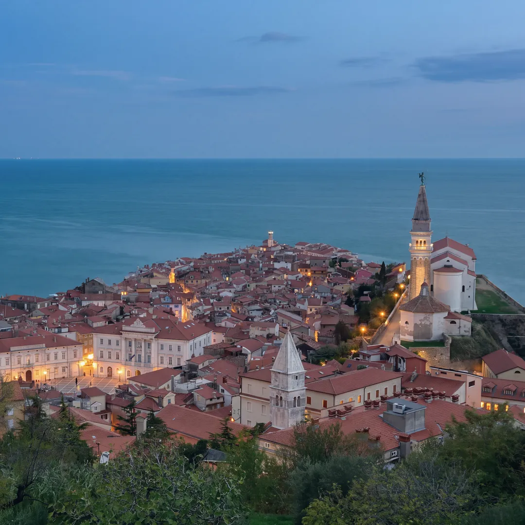 Piran from above at blue hour, from the hill of the town walls: the red roofs of the old town down to the tip, Tartini Square with the town hall and the first street lamps lit, on the right the church of St George with its lit bell tower and the octagonal baptistery, another bell tower in the foreground; beyond, the open sea.